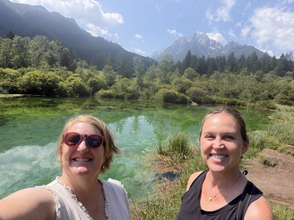 two women in front of green lake with mountains in back